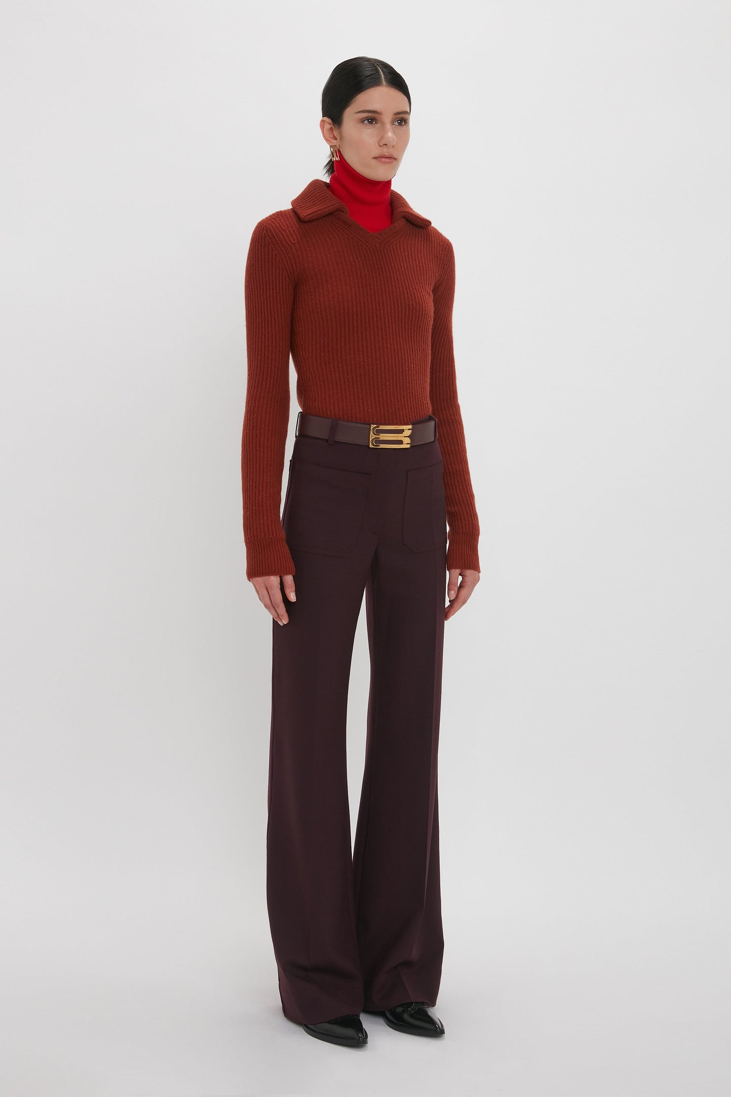 A person stands against a white background wearing a brown ribbed sweater made from recycled wool, paired with a red turtleneck and the Alina Trouser In Deep Mahogany by Victoria Beckham, cinched together with a belt featuring a gold buckle.