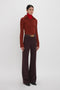 A person stands against a white background wearing a brown ribbed sweater made from recycled wool, paired with a red turtleneck and the Alina Trouser In Deep Mahogany by Victoria Beckham, cinched together with a belt featuring a gold buckle.