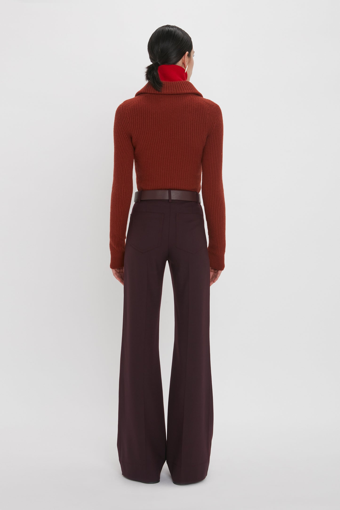 A person stands facing away, wearing a maroon ribbed turtleneck sweater, reminiscent of a Victoria Beckham Double Collared Jumper In Russet, dark high-waisted wide-leg pants, and a red scarf, against a plain white background.