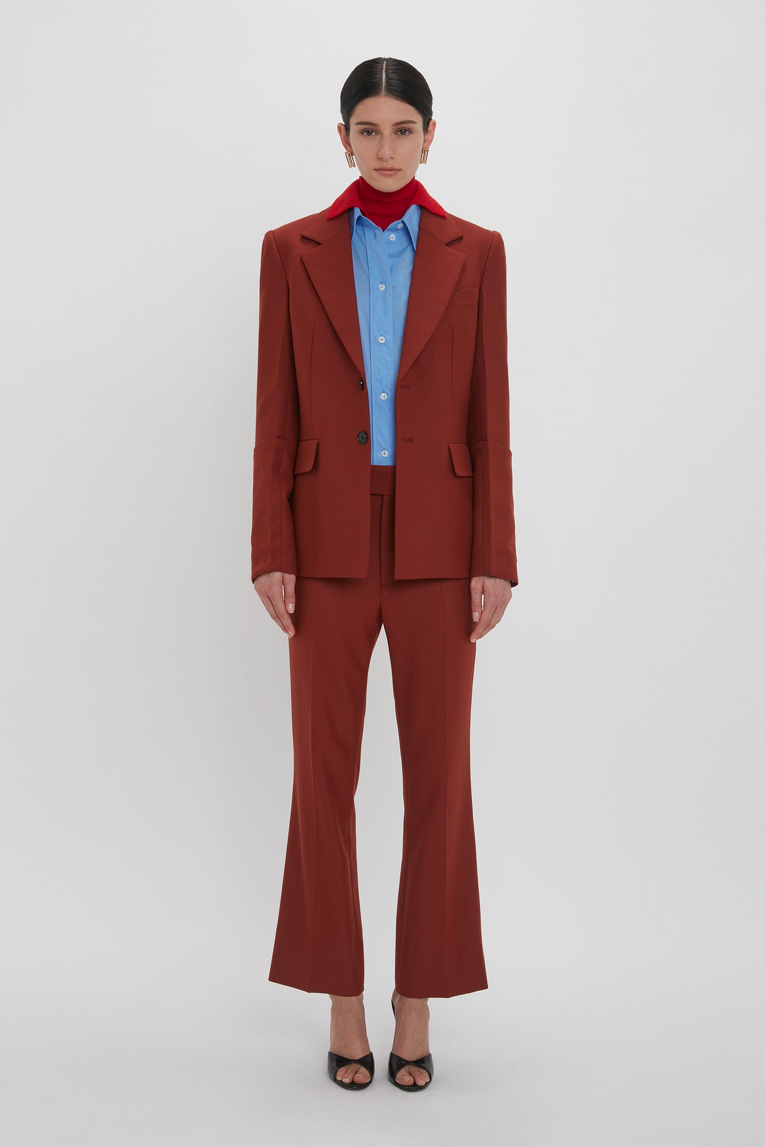 A person stands against a plain white background wearing a rust-colored suit with Wide Cropped Flare Trouser In Russet over a blue shirt, complemented by a red scarf around the neck and black heeled shoes, exuding the timeless elegance of Victoria Beckham AW24.