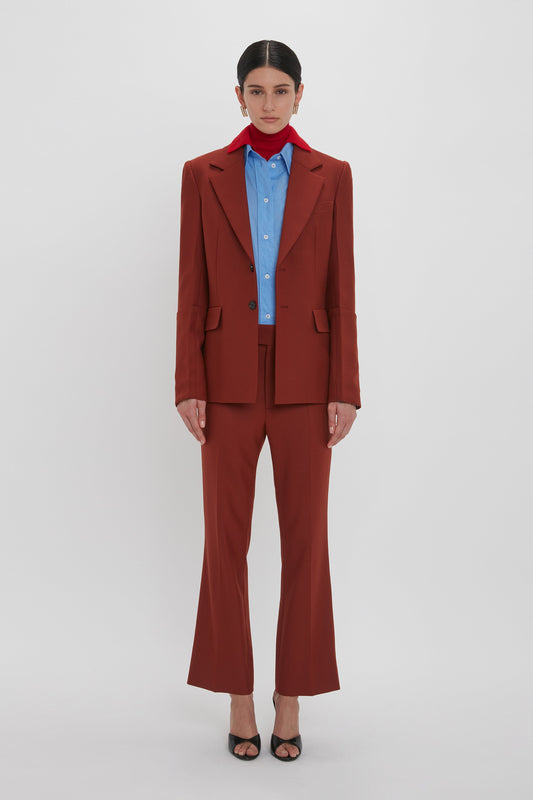 A person stands against a plain white background wearing a rust-colored suit with Wide Cropped Flare Trouser In Russet over a blue shirt, complemented by a red scarf around the neck and black heeled shoes, exuding the timeless elegance of Victoria Beckham AW24.