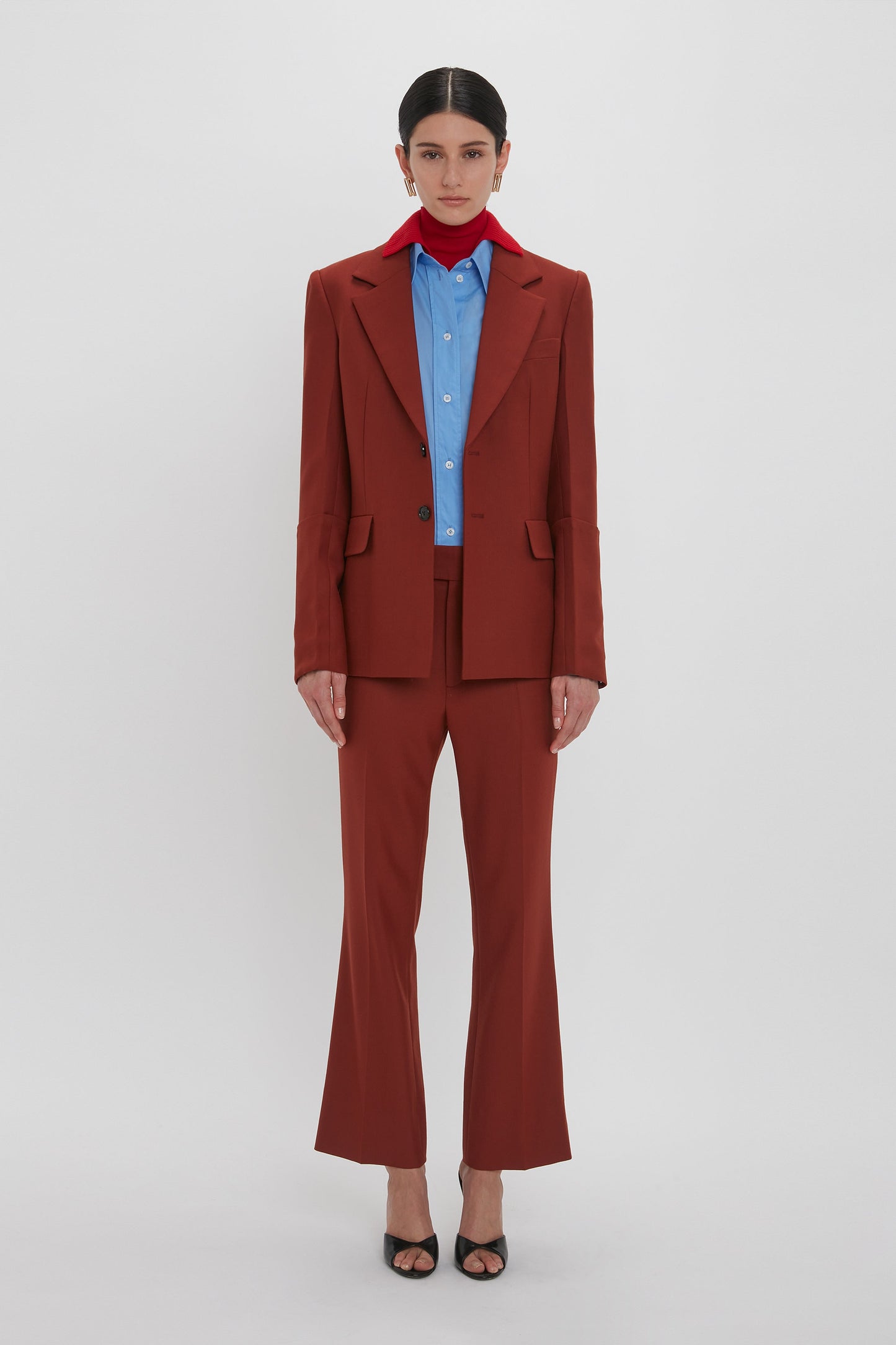 A person stands against a plain white background wearing the Victoria Beckham Sleeve Detail Patch Pocket Jacket In Russet, coupled with a blue shirt, red scarf, and black heeled sandals.
