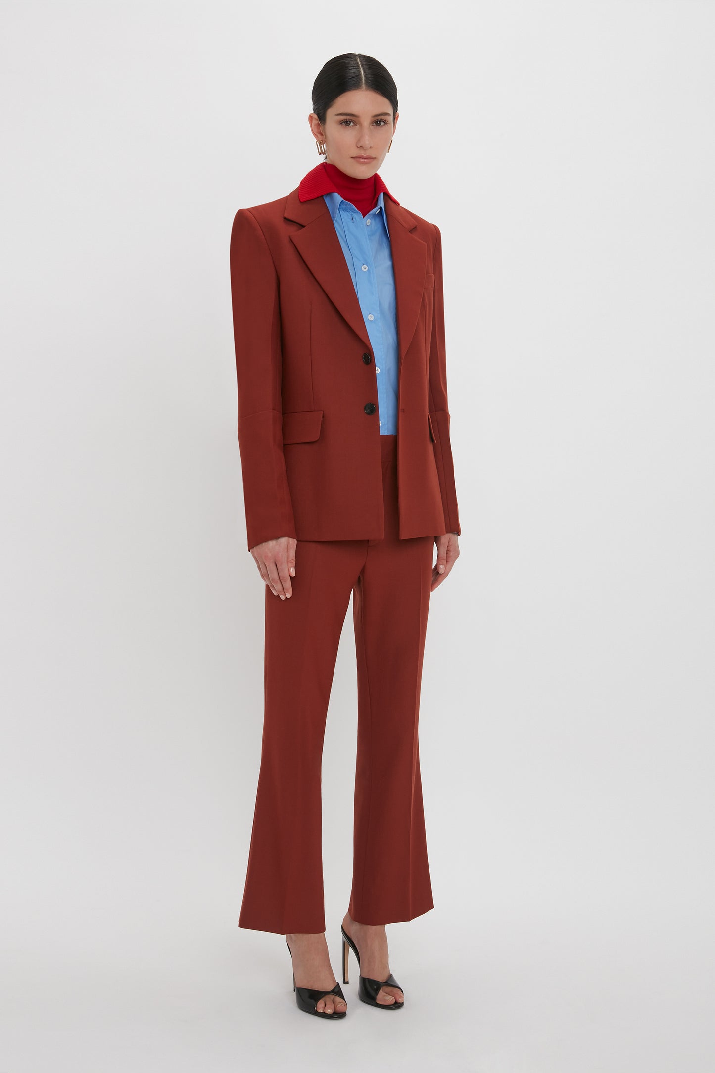 Person standing against a white background wearing the Victoria Beckham Sleeve Detail Patch Pocket Jacket in Russet with contemporary detailing, blue shirt, and red scarf. The person is also wearing black high-heeled sandals and large earrings.