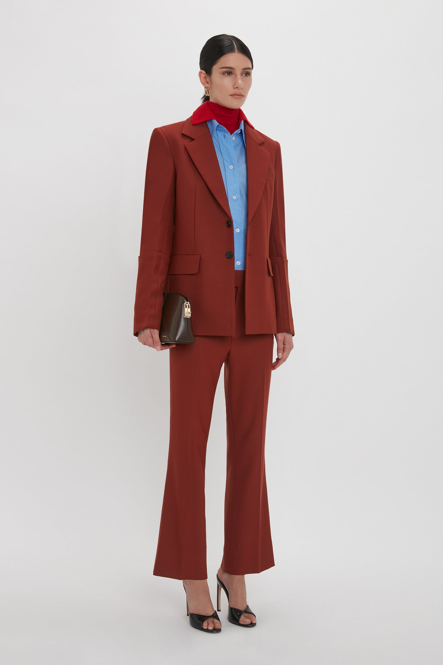 A woman in a red suit with a blue shirt and red scarf, holding a brown clutch, and wearing black heels, Victoria AW24’s signature look is complete with Victoria Beckham's Wide Cropped Flare Trouser In Russet that elevate her ensemble.