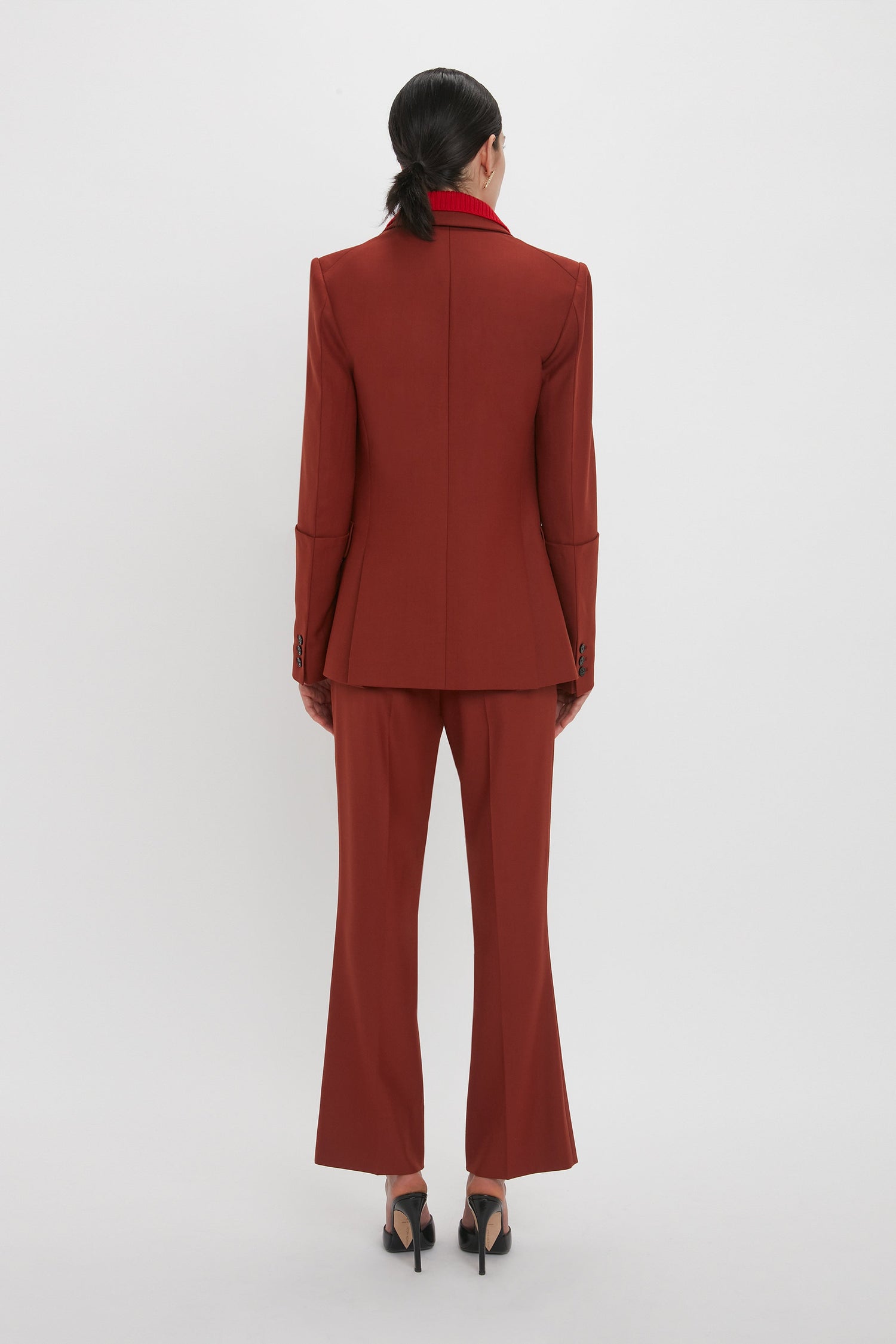 A woman with black hair is shown from the back, wearing a Sleeve Detail Patch Pocket Jacket In Russet by Victoria Beckham with matching trousers and black heels, standing against a plain white background. The contemporary detailing of her outfit adds a sophisticated touch.