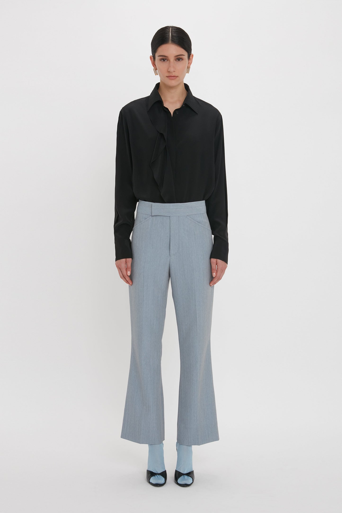 A person stands against a white background, wearing a black blouse, Victoria Beckham Exclusive Wide Cropped Flare Trouser In Marina in light grey wide-leg style with a flattering hint of ankle, and blue socks paired with black shoes.