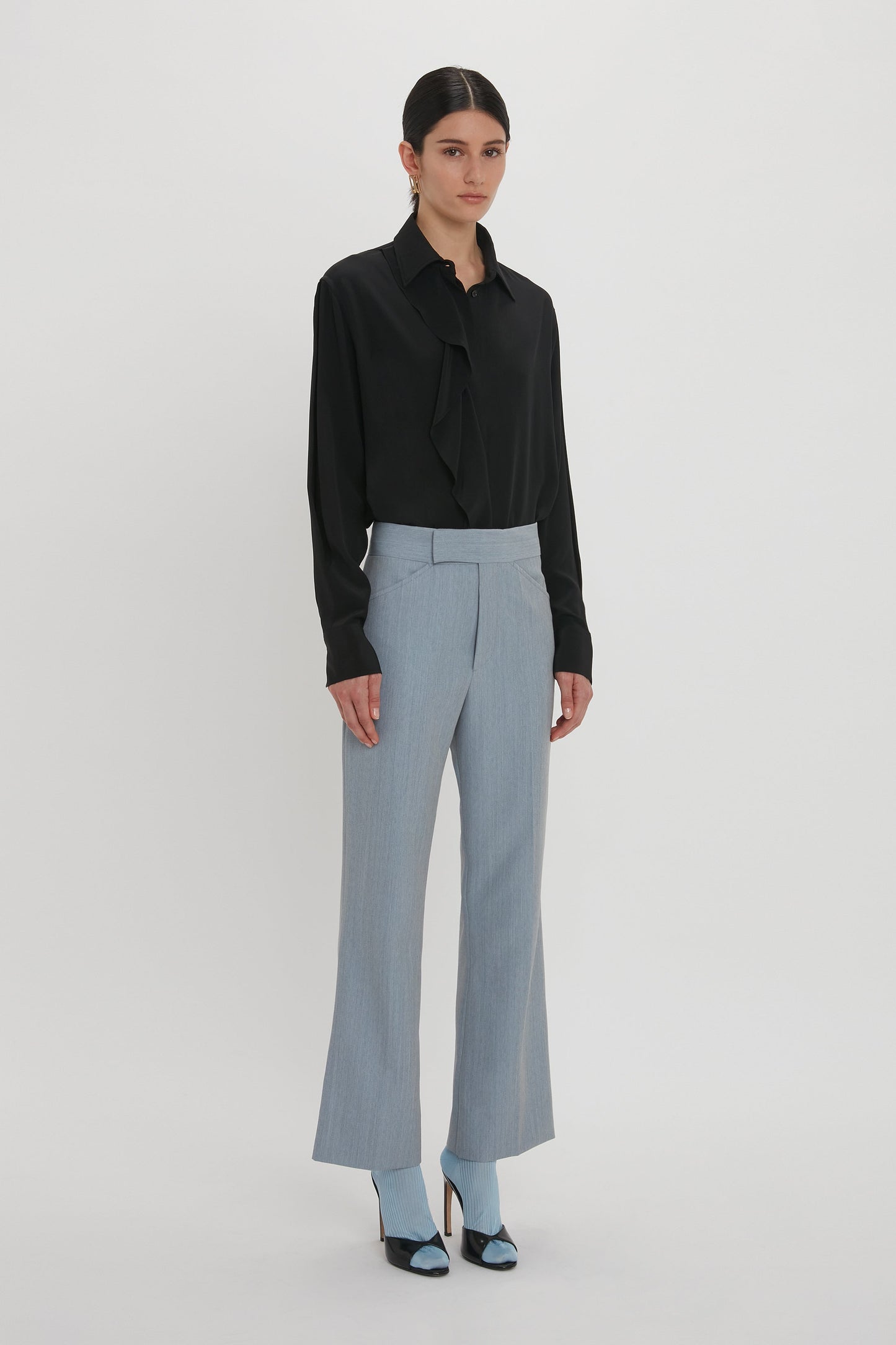 A person with dark hair is wearing a black blouse, Exclusive Wide Cropped Flare Trouser In Marina by Victoria Beckham, and blue high-heeled shoes. They are standing against a plain white background.
