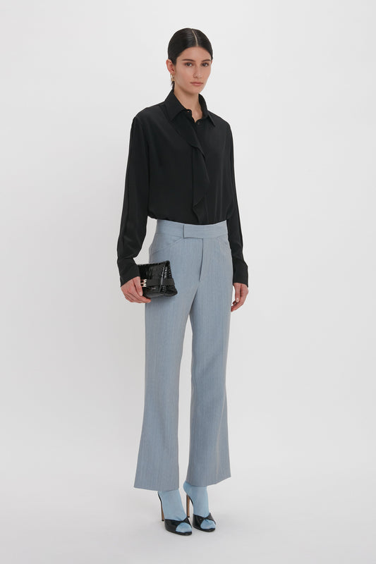 A person stands against a white background wearing a black blouse, light blue pants, and black shoes, holding a Victoria Beckham Mini B Pouch Bag In Croc Effect Black Leather.