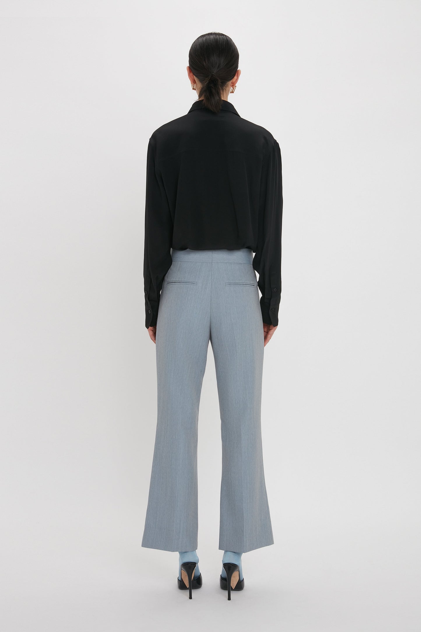 A person stands facing away, wearing a black long-sleeve shirt and Victoria Beckham Exclusive Wide Cropped Flare Trouser In Marina with a contemporary kick hem that offers a flattering hint of ankle, paired with black high heels against a plain white background.