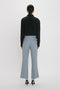 A person stands facing away, wearing a black long-sleeve shirt and Victoria Beckham Exclusive Wide Cropped Flare Trouser In Marina with a contemporary kick hem that offers a flattering hint of ankle, paired with black high heels against a plain white background.