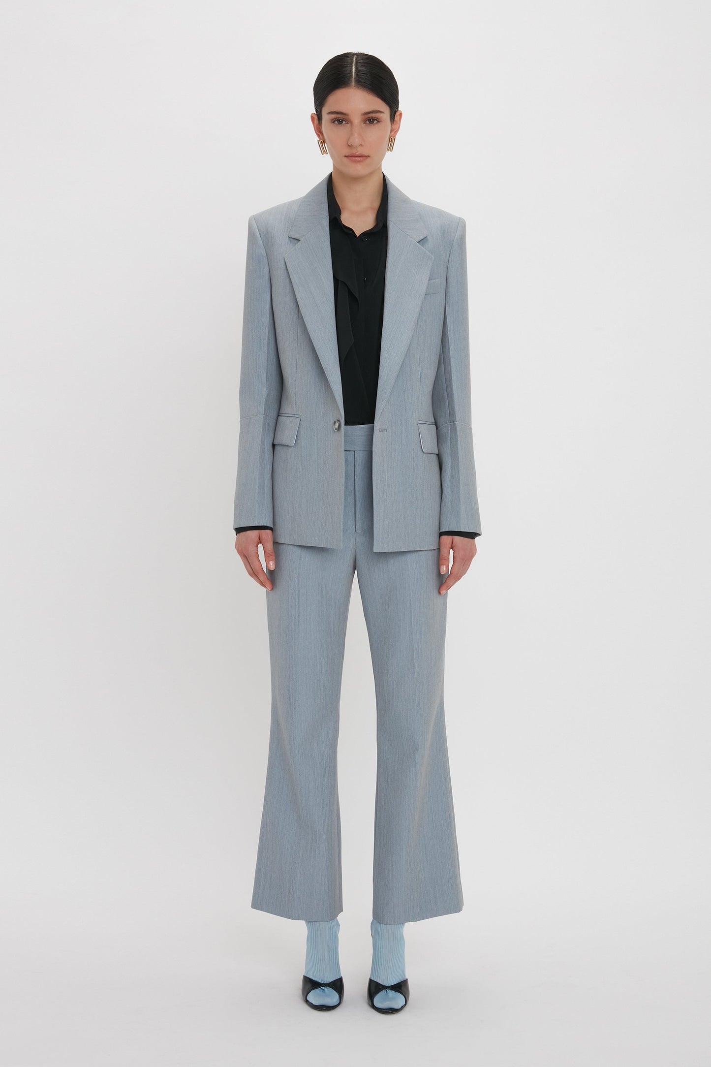 A person stands against a white background wearing the Victoria Beckham Exclusive Sleeve Detail Patch Pocket Jacket In Marina, paired with a black shirt and blue shoes.