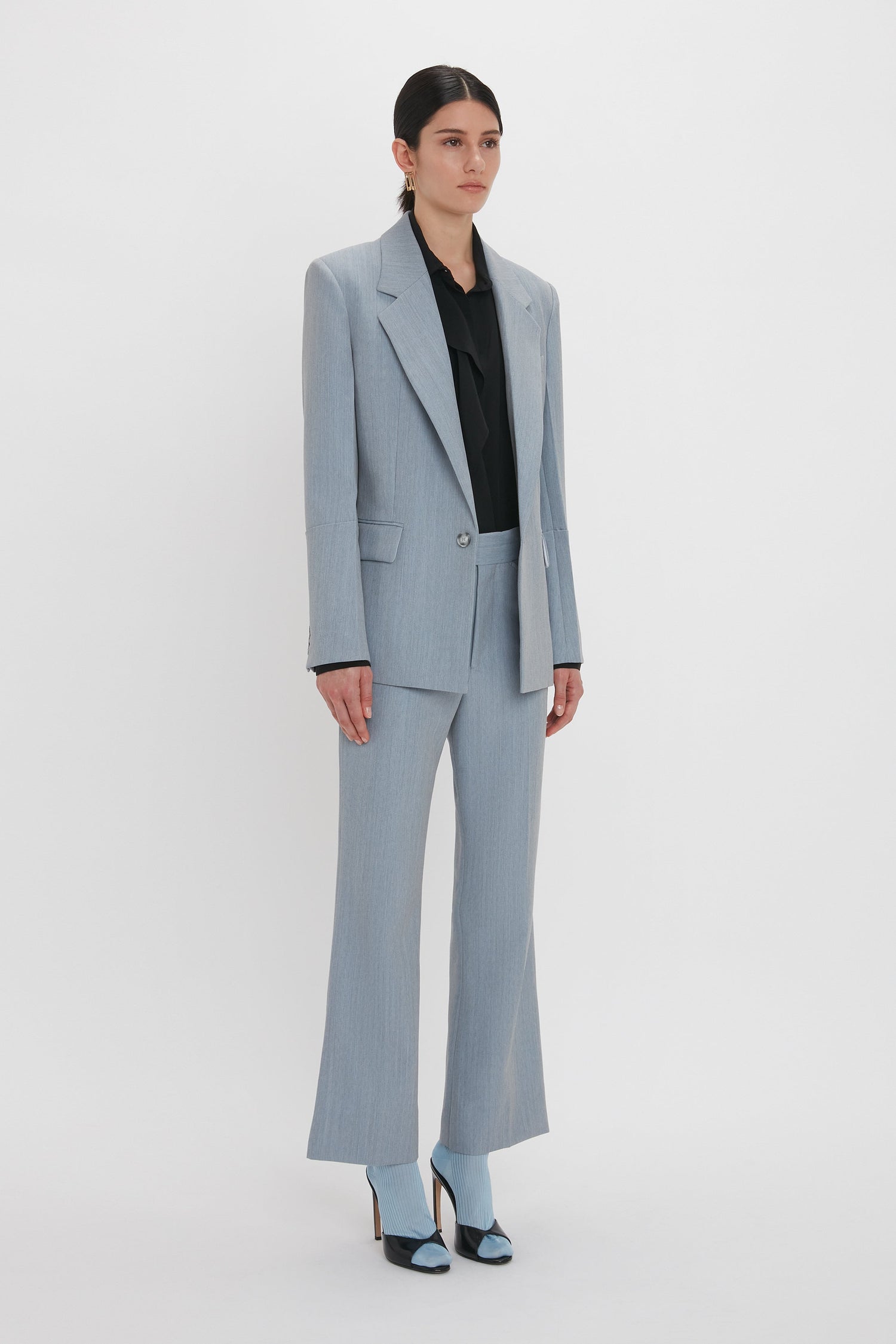 Person standing against a plain background, wearing a light gray pantsuit with a black top and coordinating light gray high-heeled shoes, completed with the Victoria Beckham Exclusive Sleeve Detail Patch Pocket Jacket In Marina featuring contemporary detailing.