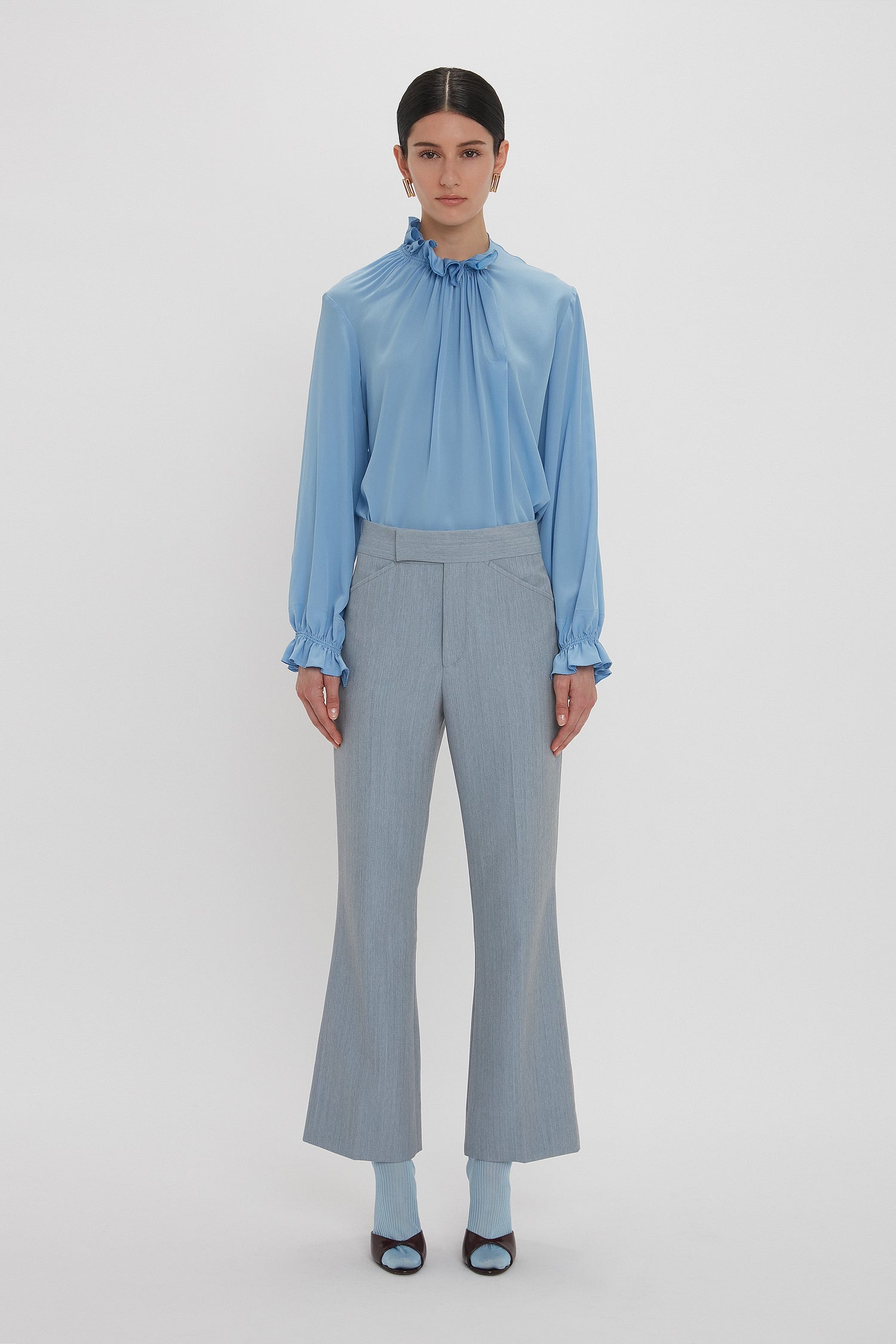 A person stands facing forward in a Victoria Beckham Exclusive Ruffle Neck Blouse In Cornflower Blue and gray pants with flared legs, set against a plain white background.