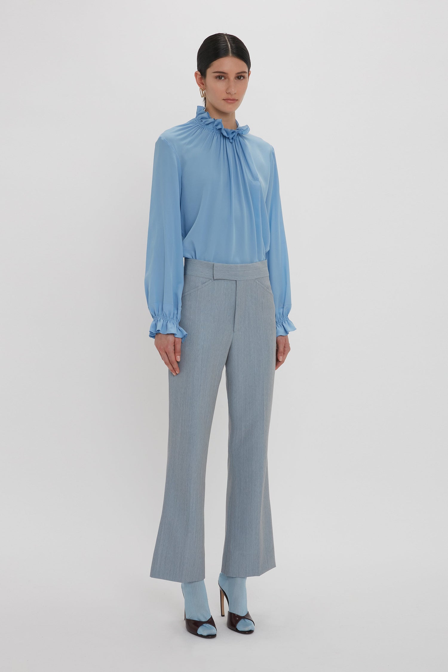 A woman stands against a plain white background, wearing the Victoria Beckham Exclusive Ruffle Neck Blouse In Cornflower Blue made from crepe de chine fabric, gray high-waisted pants, and matching cornflower blue shoes with open toes.