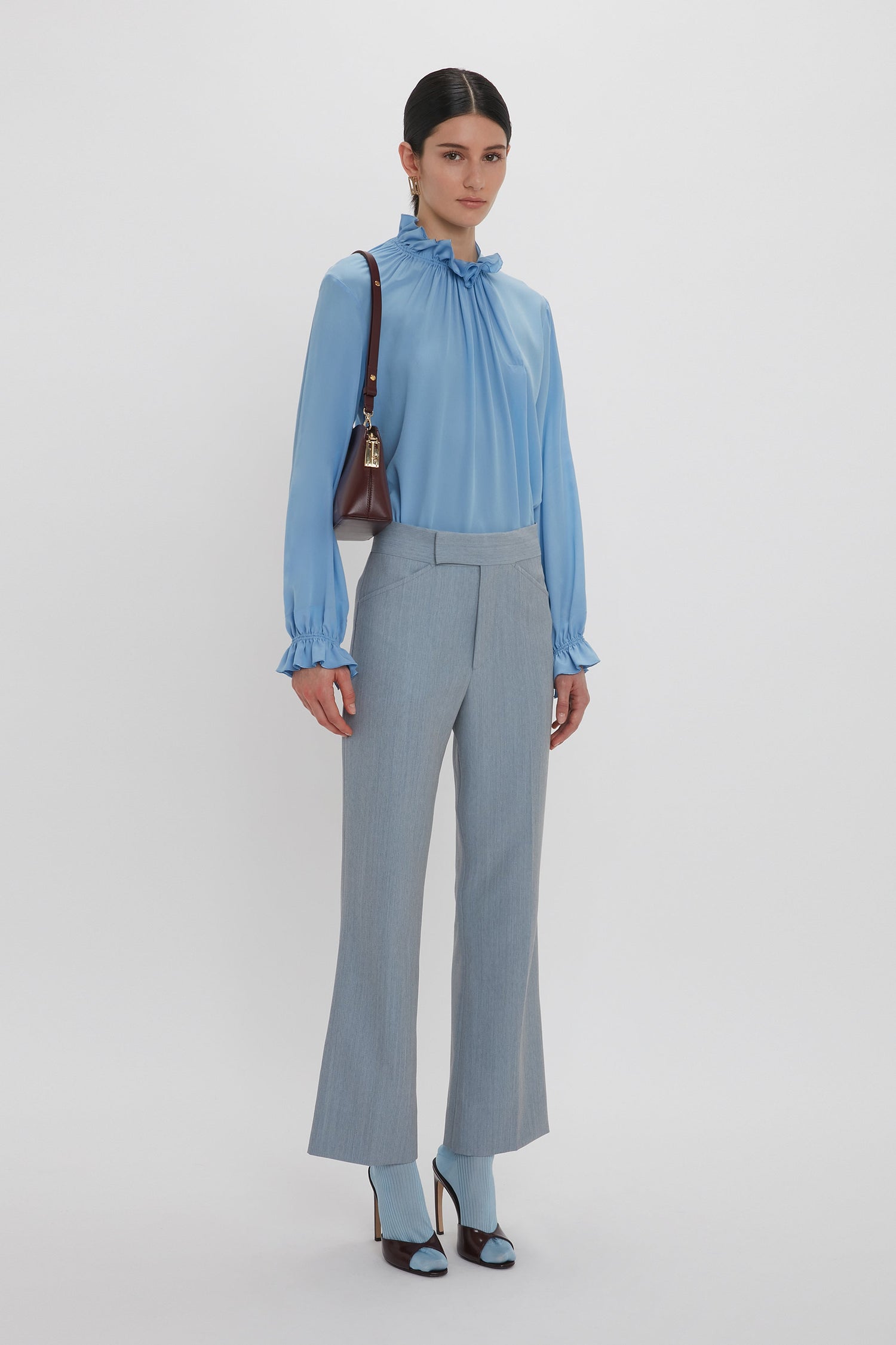A person wearing a Victoria Beckham Exclusive Ruffle Neck Blouse in Cornflower Blue, gray trousers, and high-heeled shoes stands against a white background, carrying a small brown handbag over their shoulder.