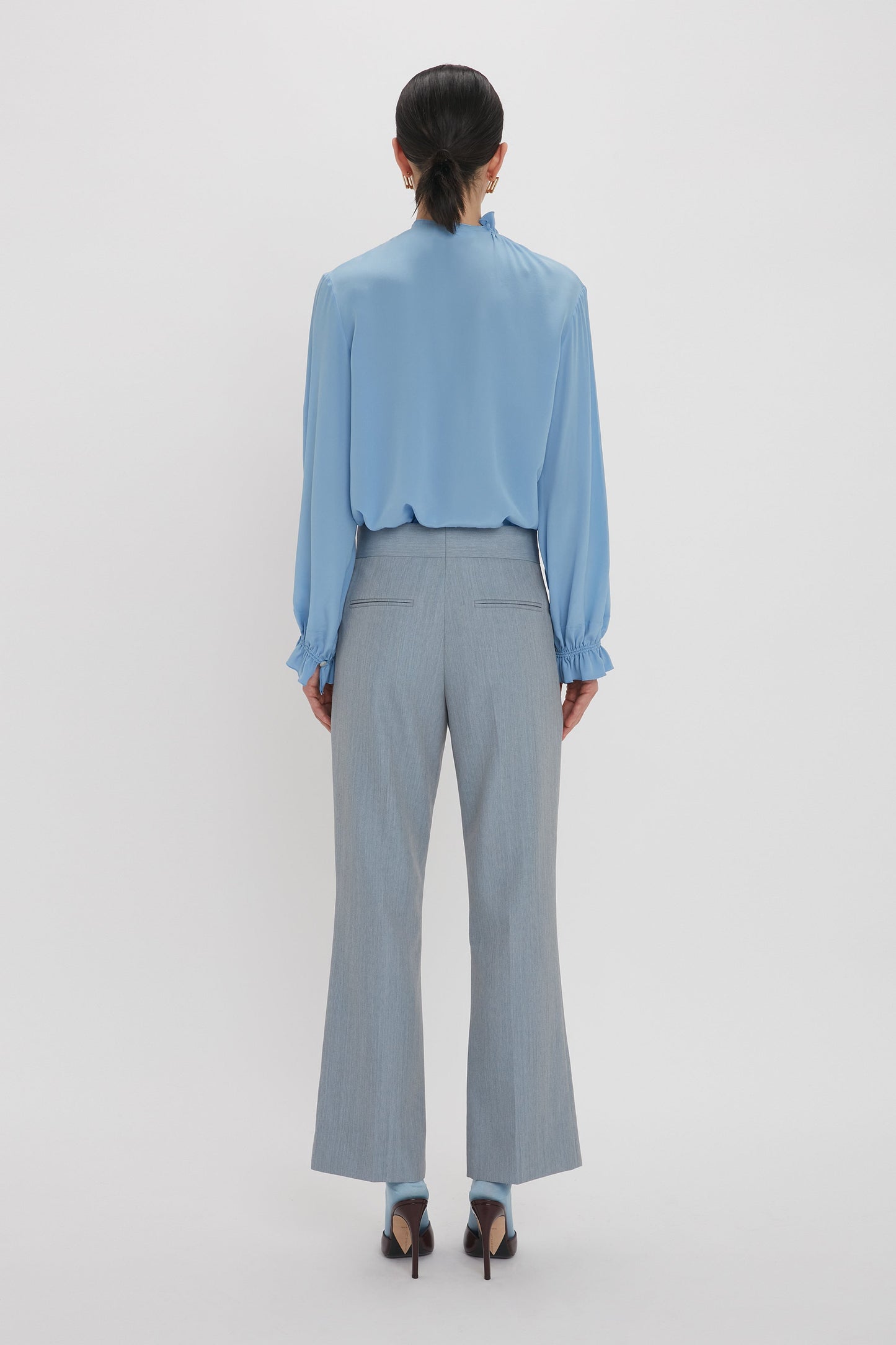 A person with dark hair is standing facing away, wearing the Victoria Beckham Exclusive Ruffle Neck Blouse In Cornflower Blue and grey high-waisted trousers with a slight flare at the bottom, paired with dark heels.
