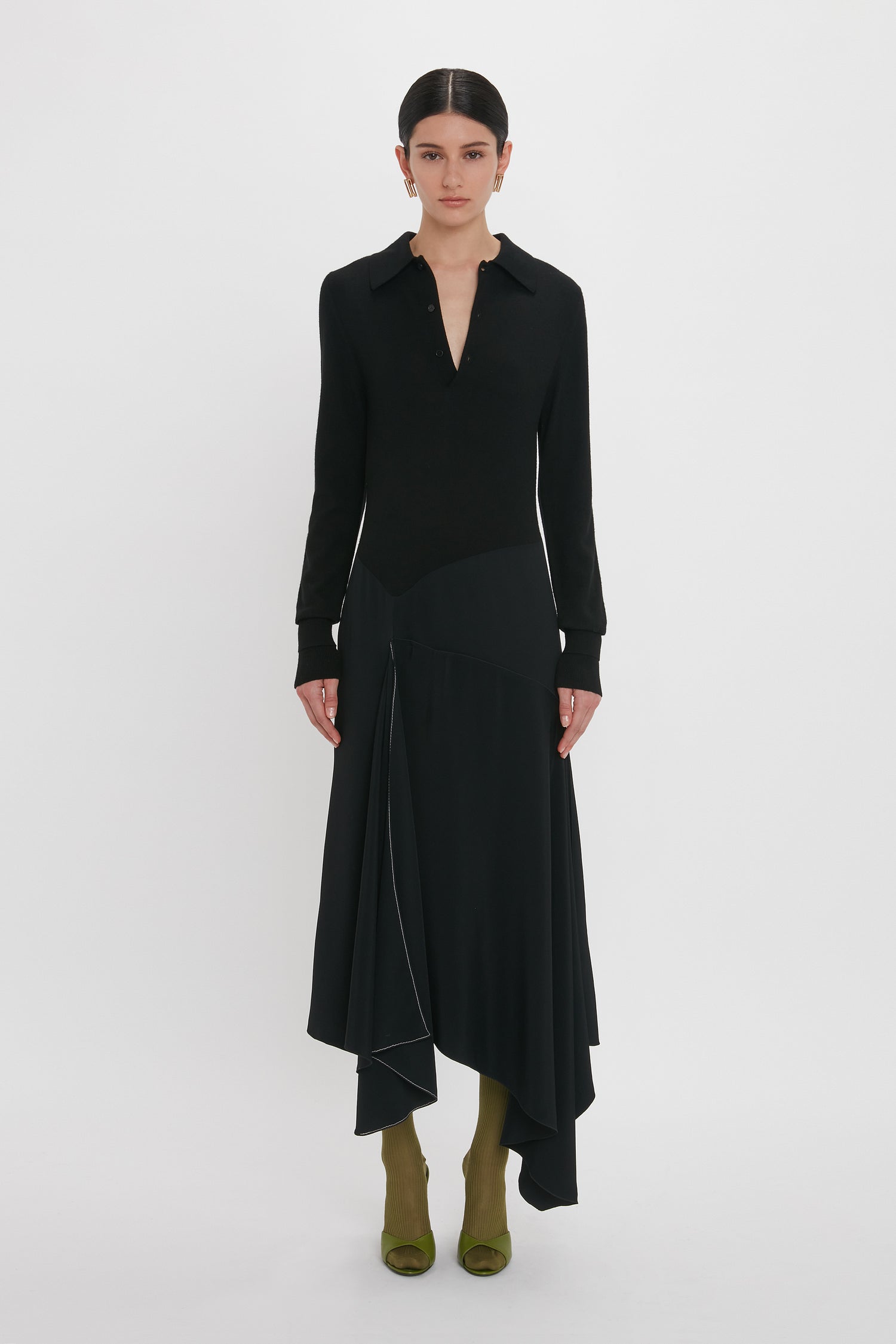 A person stands against a white background wearing a Victoria Beckham Henley Shirt Dress In Black with an asymmetrical hem and green shoes.