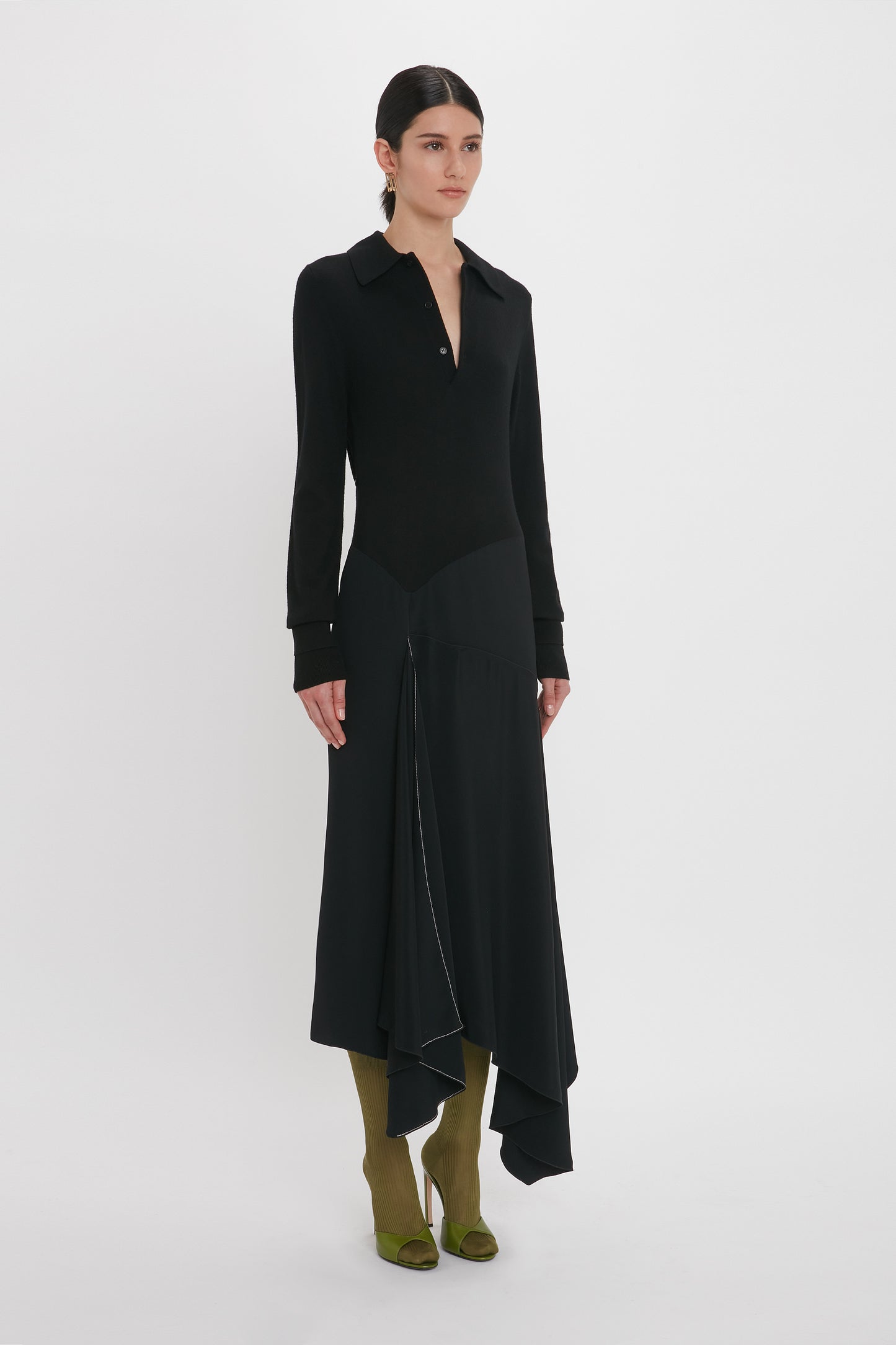 A person wearing a long black Henley Shirt Dress In Black by Victoria Beckham with a collar, long sleeves, and an asymmetric waist seam, paired with green knee-high boots, standing against a plain white background.