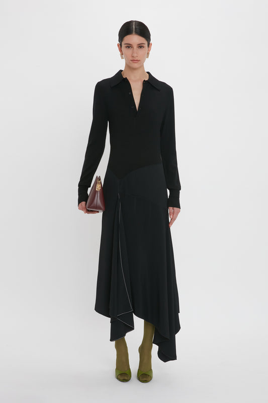 A person stands against a white background wearing a Victoria Beckham Henley Shirt Dress In Black with an asymmetric waist seam and olive-green shoes, holding a small maroon purse.