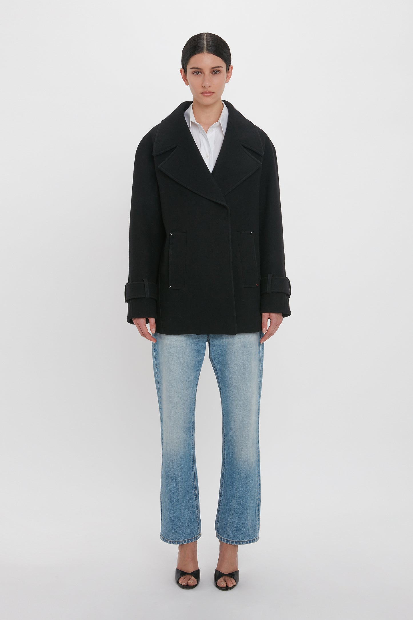 A person stands against a plain white background wearing an Oversized Pea Coat In Black by Victoria Beckham, along with a white shirt, blue jeans, and black open-toe shoes. They have their hands by their sides and are looking directly at the camera.