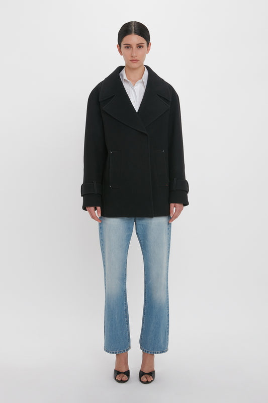 A person stands against a plain white background wearing an Oversized Pea Coat In Black by Victoria Beckham, along with a white shirt, blue jeans, and black open-toe shoes. They have their hands by their sides and are looking directly at the camera.