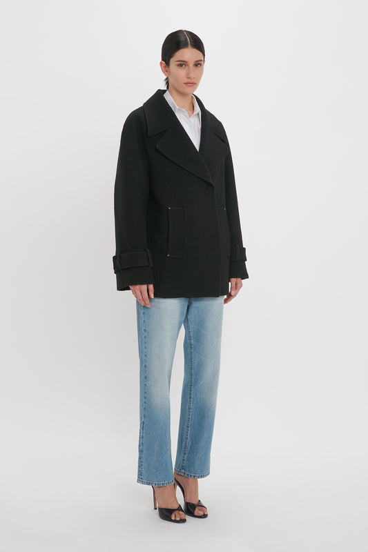 A person stands against a plain background wearing an Oversized Pea Coat In Black by Victoria Beckham, light blue jeans, and black heels.