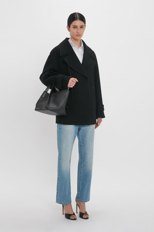 A person stands against a white background wearing an Oversized Pea Coat In Black by Victoria Beckham, blue jeans, black heels, and holding a black handbag.