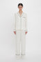 A person stands against a plain white background wearing the Victoria Beckham VB Monogram Pyjama Sleep Set In Ivory with black piping. The elegant loungewear includes a long-sleeved button-up shirt and loose-fitting pants, tailored from 100% silk for ultimate comfort.