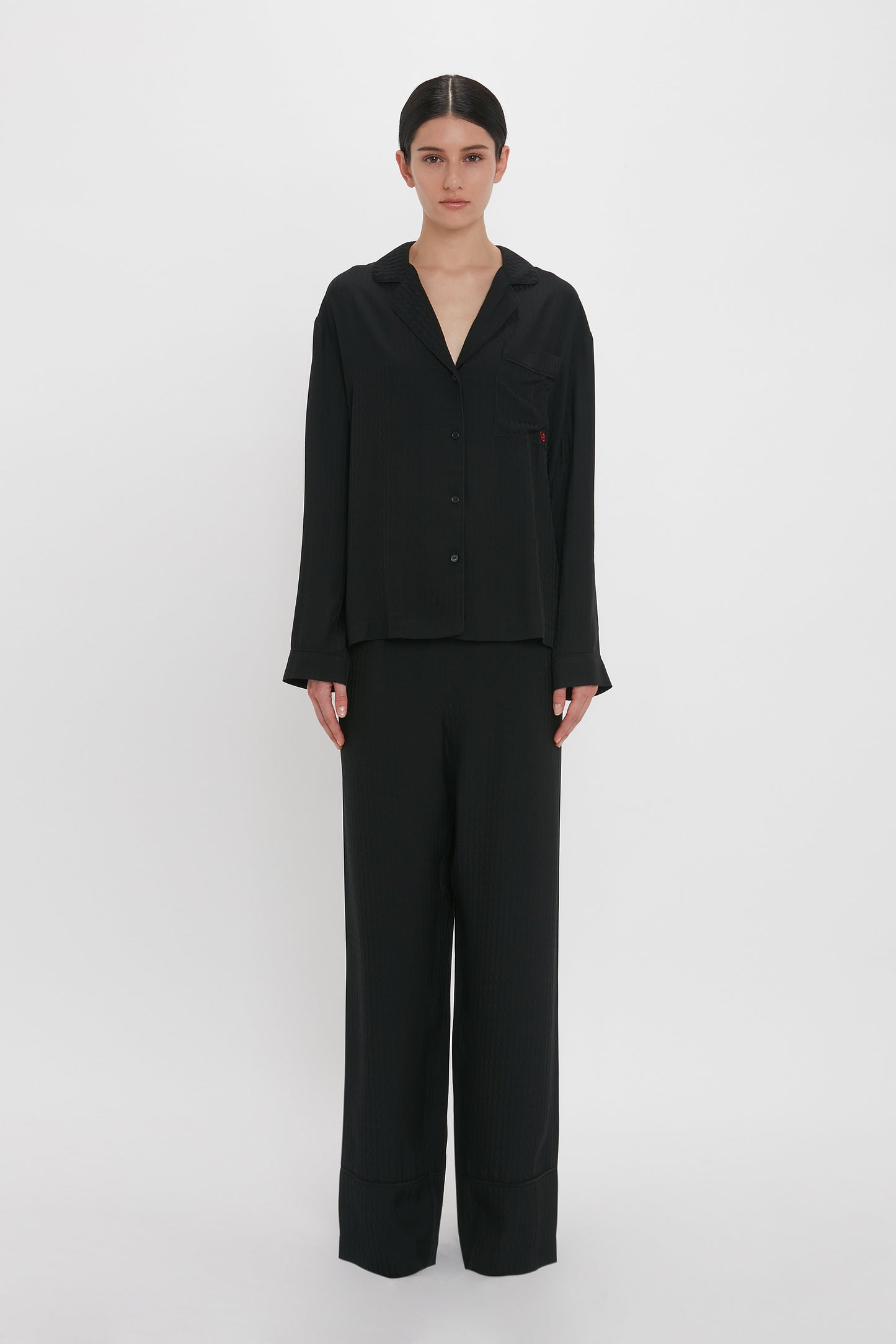 Person standing against a plain white background, wearing a black long-sleeve button-up shirt and matching black pants, reminiscent of the luxurious VB Monogram Pyjama Sleep Set In Black by Victoria Beckham loungewear style.