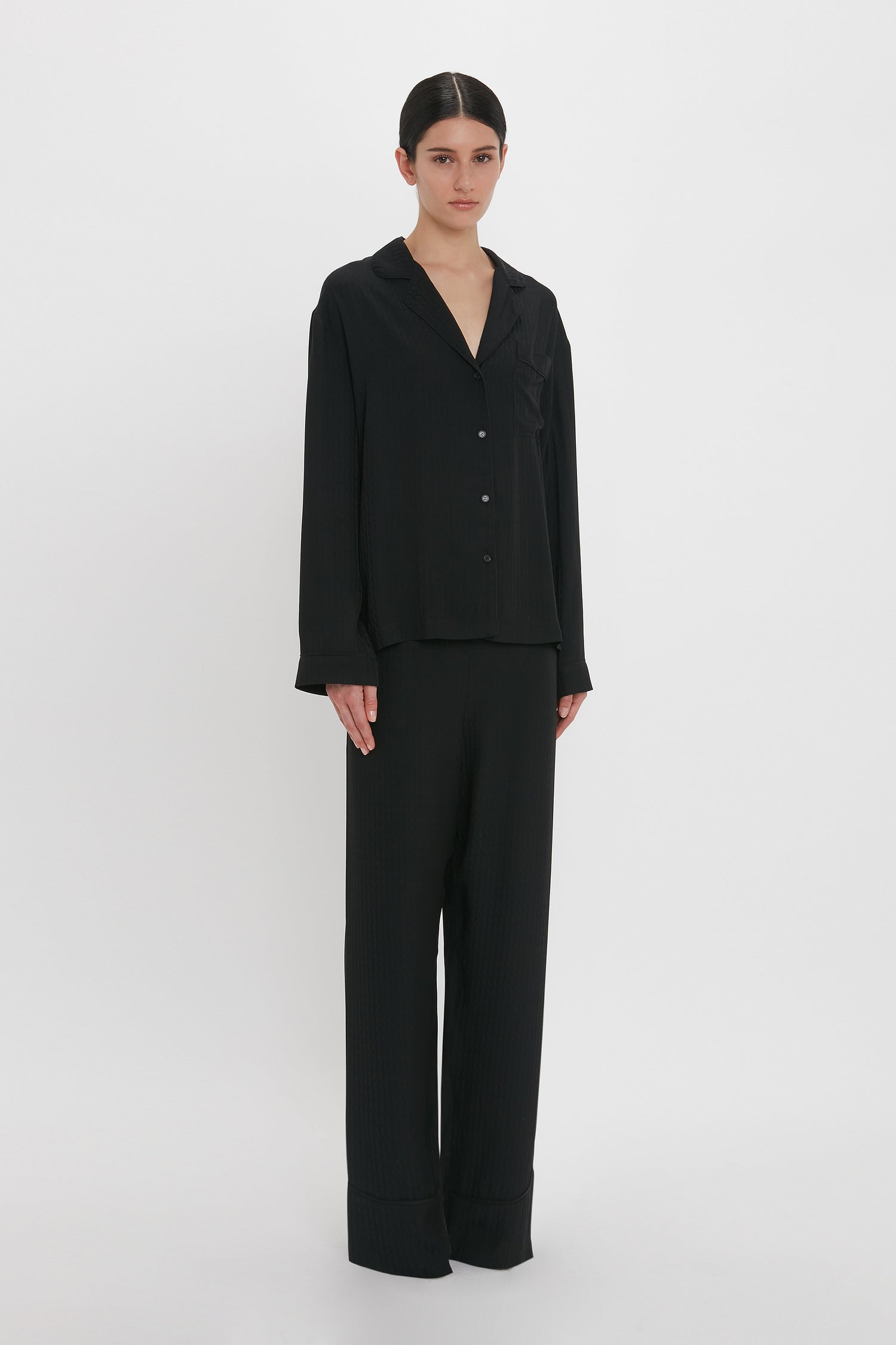 Person standing against a white background, wearing the Victoria Beckham VB Monogram Pyjama Sleep Set In Black, a luxurious black long-sleeve button-up shirt and matching loose-fitting pants, reminiscent of an elegant silk pyjama sleep set.