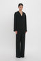 Person standing against a white background, wearing the Victoria Beckham VB Monogram Pyjama Sleep Set In Black, a luxurious black long-sleeve button-up shirt and matching loose-fitting pants, reminiscent of an elegant silk pyjama sleep set.