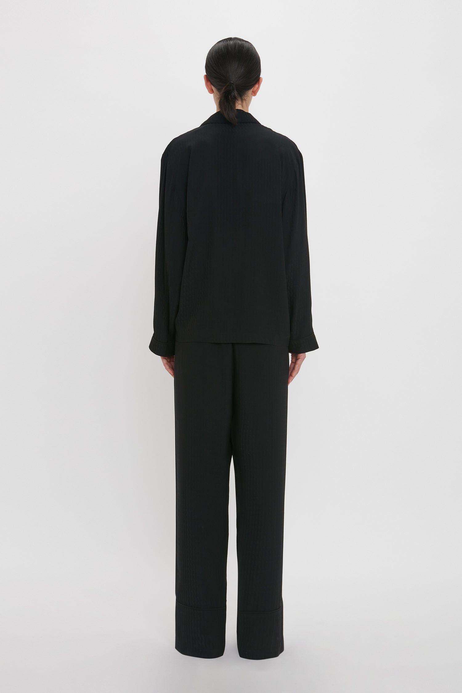 Rear view of a person with dark hair wearing a luxurious VB Monogram Pyjama Sleep Set In Black by Victoria Beckham, complete with a long-sleeved black shirt and black pants, standing against a plain white background.