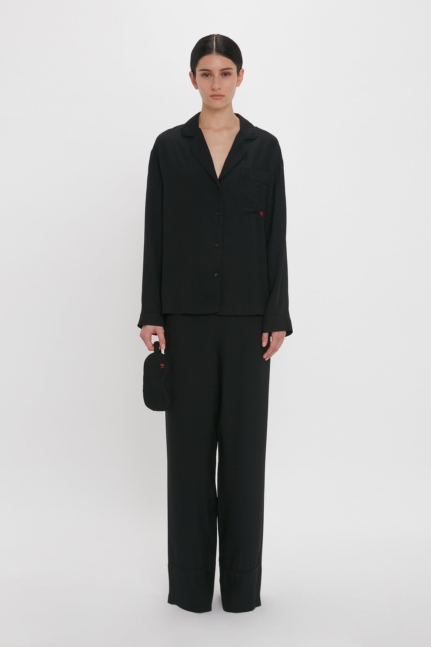 A person with dark hair tied back wears a black button-up top and matching black pants, holding a small black pouch at their side, reminiscent of luxurious loungewear, the VB Monogram Pyjama Sleep Set In Black by Victoria Beckham. The background is plain white.