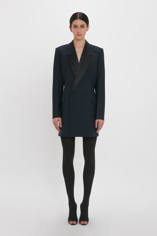 A person stands against a white background wearing a black tailored blazer dress, black tights, and black open-toe shoes. This chic ensemble is perfect for evening wear and exudes the sophistication of the Victoria Beckham Exclusive Fold Shoulder Detail Dress In Midnight.