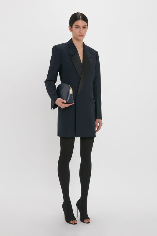 Person wearing an elegant Victoria Beckham Exclusive Fold Shoulder Detail Dress In Midnight, black tights, and black high heels, holding a small black purse.