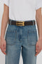 Person wearing a white t-shirt, blue jeans with front patch pockets, and a Jumbo Frame Belt In Espresso Croc Embossed Calf Leather by Victoria Beckham.