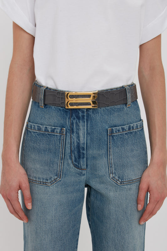 A person wearing a white shirt and blue jeans with a Victoria Beckham Jumbo Frame Belt In Slate Grey Croc Embossed Calf Leather featuring a gold buckle.