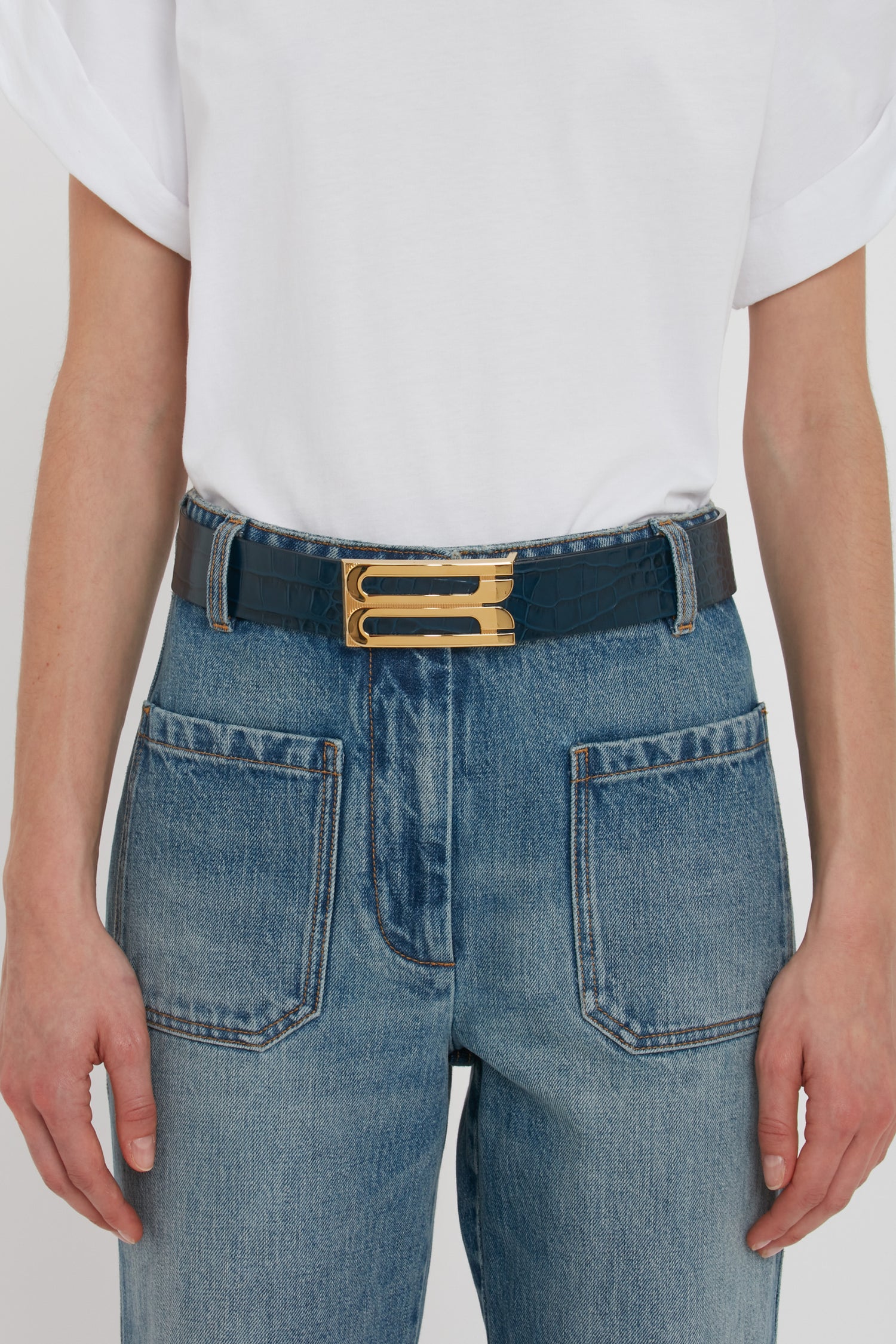 A person wearing a white t-shirt tucked into blue jeans, accessorized with a Jumbo Frame Belt In Midnight Blue Croc Embossed Calf Leather crafted from croc-embossed calf leather and featuring luxury gold hardware by Victoria Beckham.