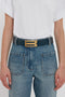 A person wearing a white t-shirt tucked into blue jeans, accessorized with a Jumbo Frame Belt In Midnight Blue Croc Embossed Calf Leather crafted from croc-embossed calf leather and featuring luxury gold hardware by Victoria Beckham.