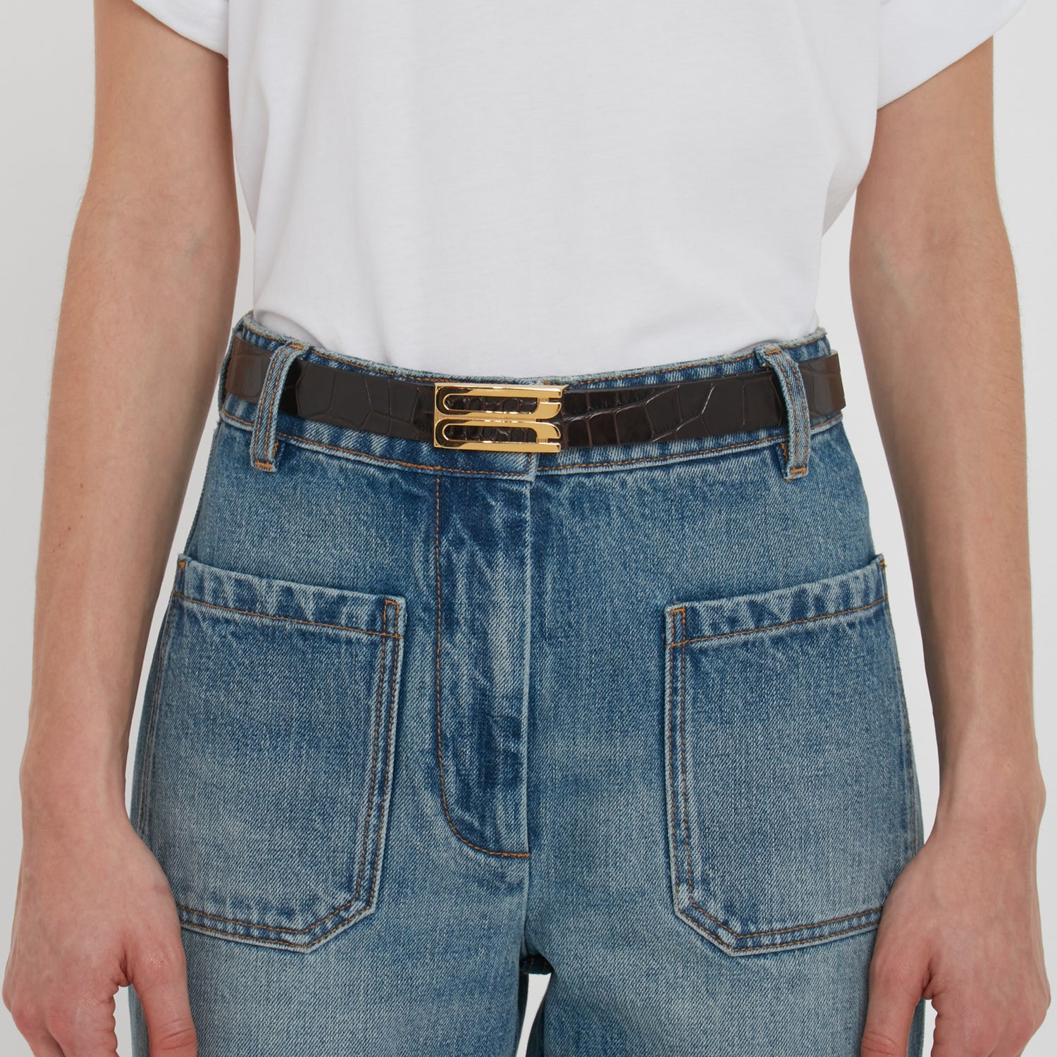 Person wears a white shirt tucked into high-waisted blue jeans with large front pockets and a black Victoria Beckham Frame Belt In Espresso Croc Embossed Calf Leather with gold hardware.