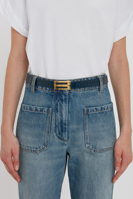 Person wearing a white t-shirt tucked into high-waisted blue jeans with large front pockets and a Victoria Beckham Frame Belt In Midnight Blue Croc Embossed Calf Leather featuring gold hardware.