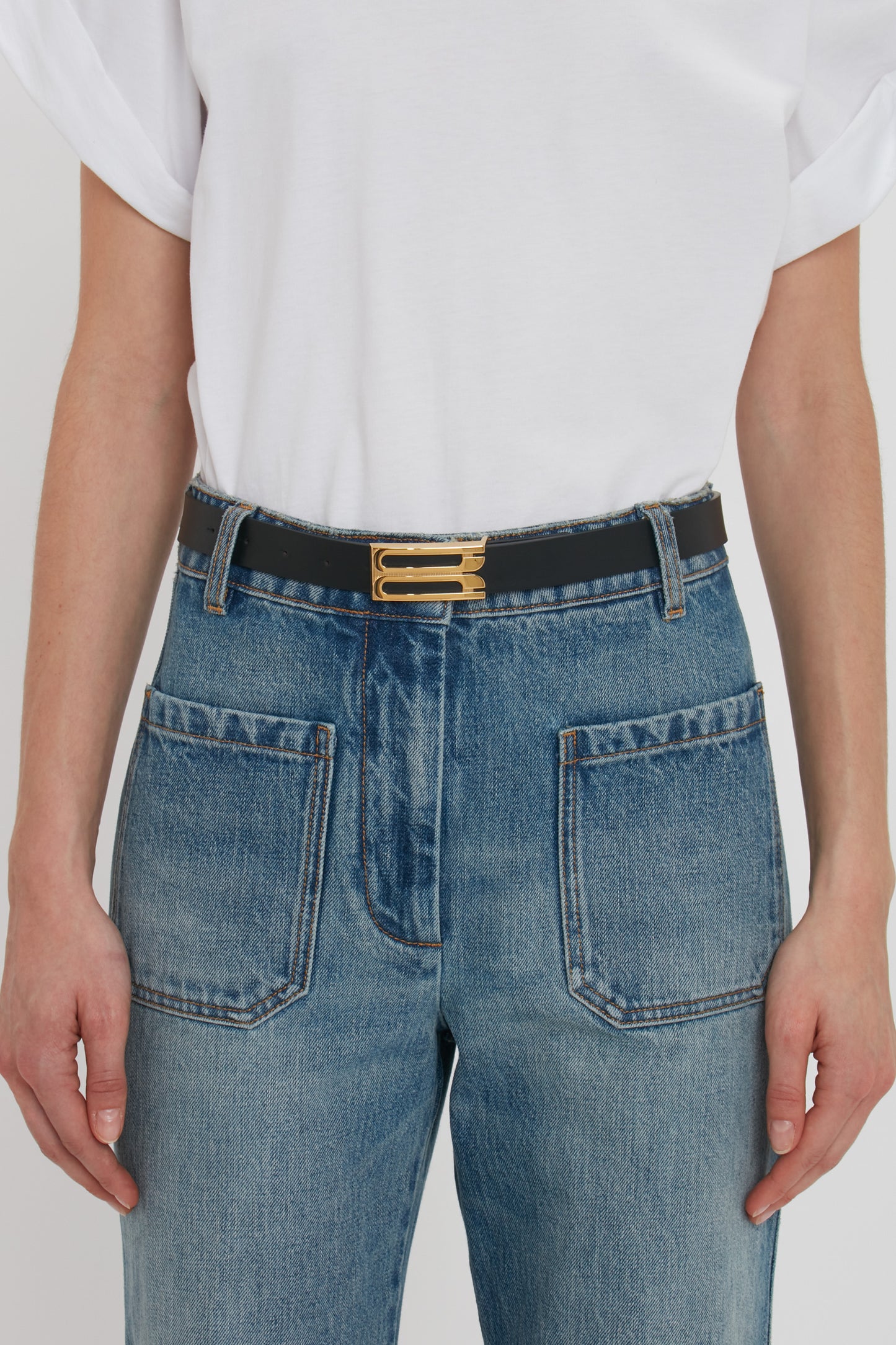 A person wearing a white shirt tucked into high-waisted blue jeans with large front pockets, and a fashion-forward Victoria Beckham Frame Belt In Black Leather.