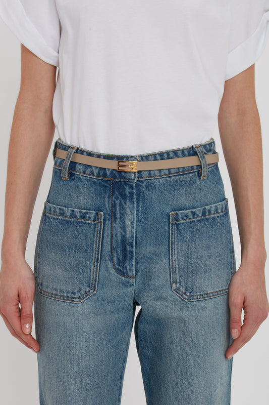 A person wearing a white t-shirt and high-waisted blue jeans with large front pockets, accessorized with a beige Exclusive Micro Frame Belt In Beige Leather featuring gold hardware by Victoria Beckham.