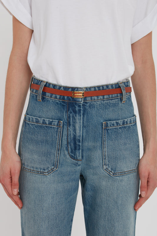 Person wearing a white shirt tucked into high-waisted blue jeans with front pockets, secured with the Exclusive Micro Frame Belt In Tan Leather by Victoria Beckham, featuring elegant gold hardware.