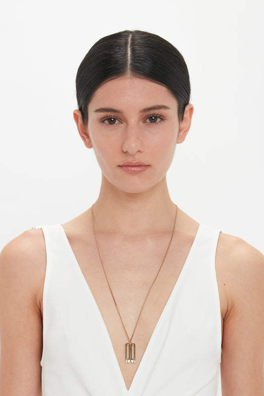 A person with dark hair in a middle part wears a white sleeveless top and a long necklace featuring the Exclusive Frame Necklace In Gold by Victoria Beckham against a plain white background, reminiscent of Victoria Beckham accessories.
