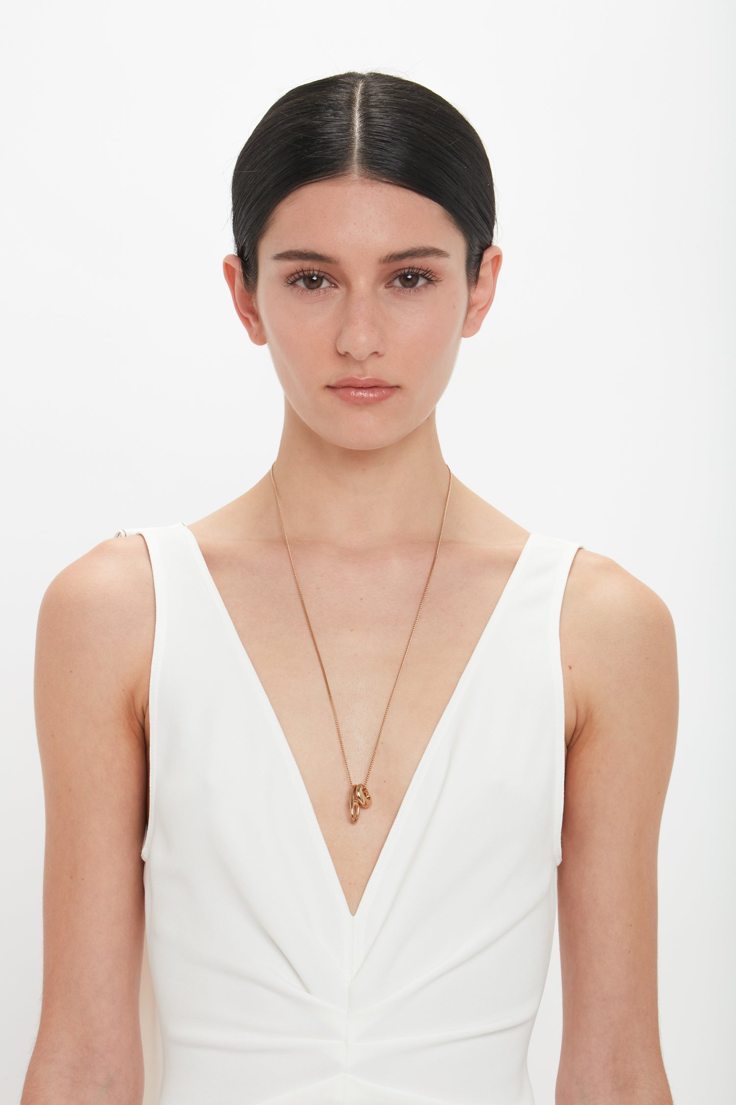 A person with dark hair tied back wears a white V-neck outfit and a Victoria Beckham Exclusive Abstract Charm Necklace In Light Gold with a T-bar closure, standing against a plain light gold background.