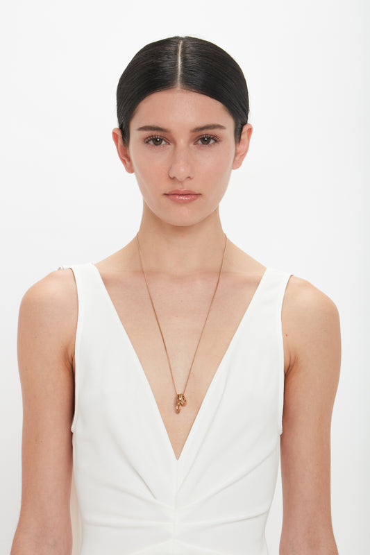 A person with dark hair tied back wears a white V-neck outfit and a Victoria Beckham Exclusive Abstract Charm Necklace In Light Gold with a T-bar closure, standing against a plain light gold background.