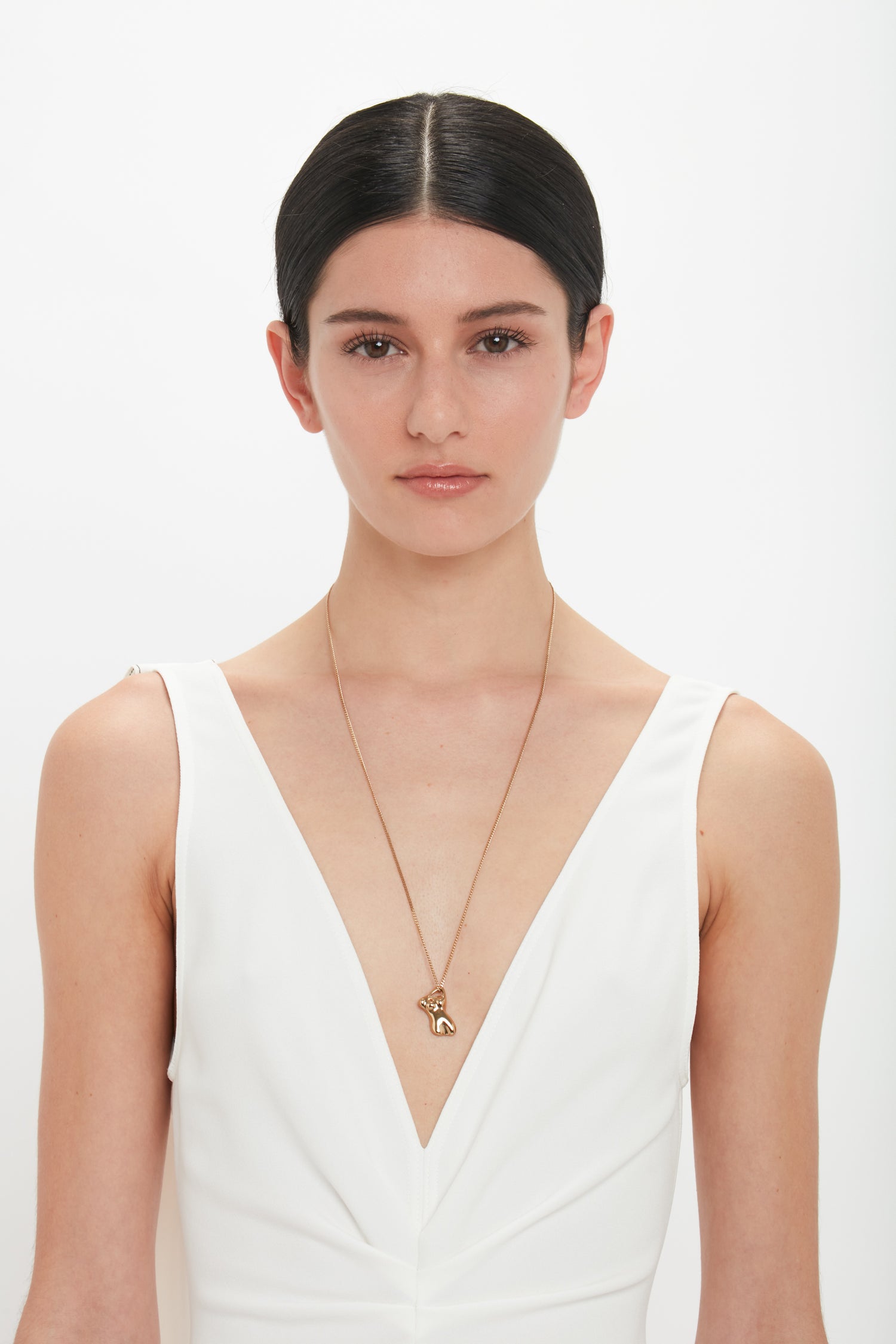 A woman with dark hair wearing a white sleeveless top and a long, light gold Exclusive Body Charm Necklace In Light Gold by Victoria Beckham, gazes directly at the camera against a plain background.