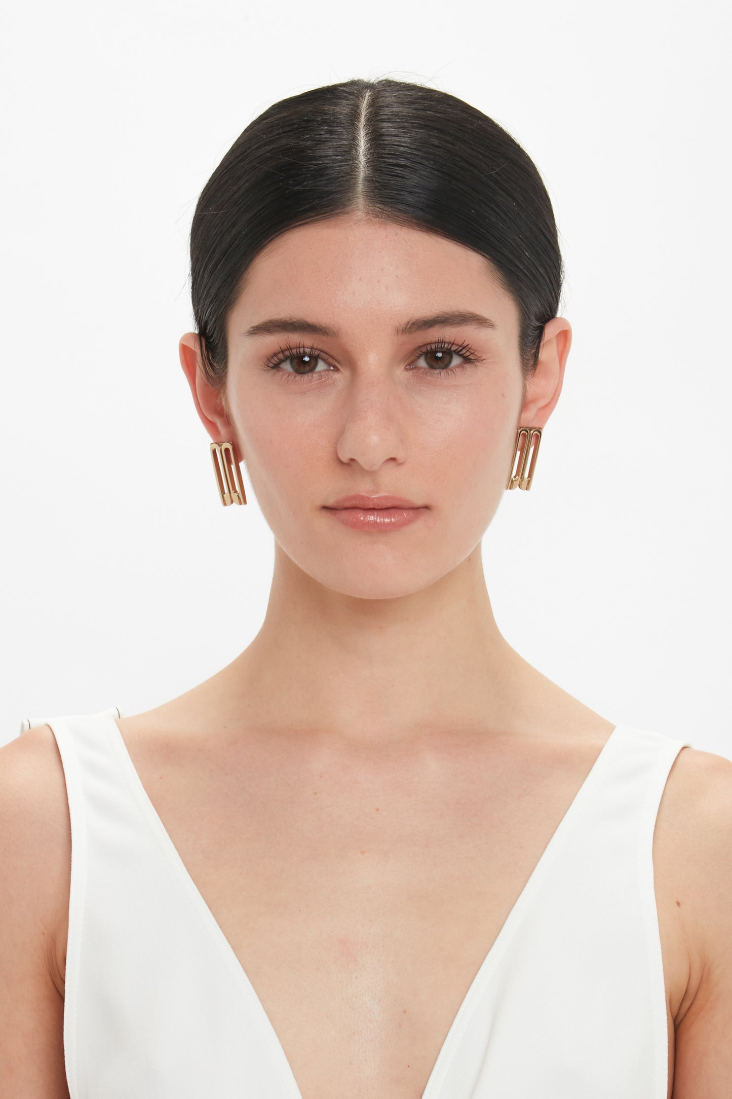 A person with dark hair tied back, wearing Victoria Beckham Exclusive Frame Stud Earrings In Gold and a white top, is looking straight ahead with a neutral expression.
