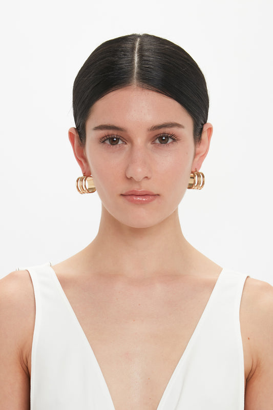 A woman with dark hair pulled back, wearing Victoria Beckham Exclusive Frame Hoop Earrings In Gold and a white sleeveless top, stares directly at the camera.